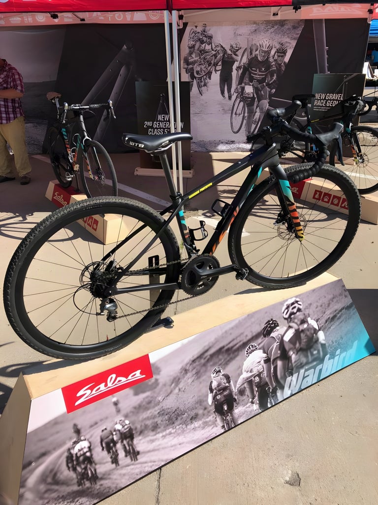 Black Salsa Warbird V4 prototype with drop bars and 700c wheels on a plinth in front of a Warbird banner at Dirty Kanza.