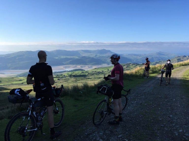 TINAT riders pause beside loaded bikes on a hillside track overlooking the estuary and hills towards Barmouth.