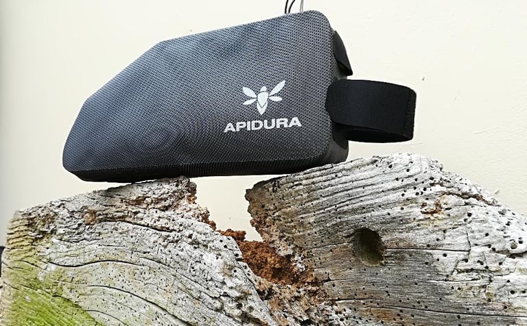 Apidura Expedition top tube bag in grey with a Velcro strap extended, balanced on a weathered log.