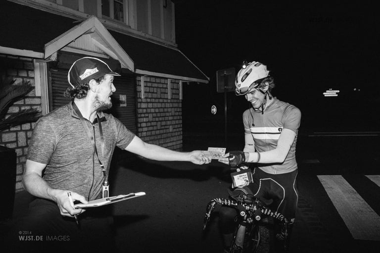 Josh Ibbett grins as Mike Hall hands him a stamped brevet card at night during the 2014 Transcontinental Race.