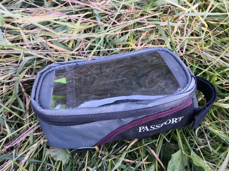 Closed Passport top-tube bag with clear window for a phone, purple trim and logo, lying on grass.