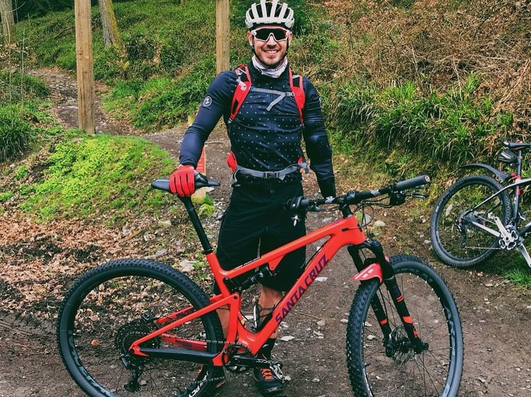 Jaco van Gass, WWTW ambassador, smiling beside a red Santa Cruz mountain bike on a woodland trail.