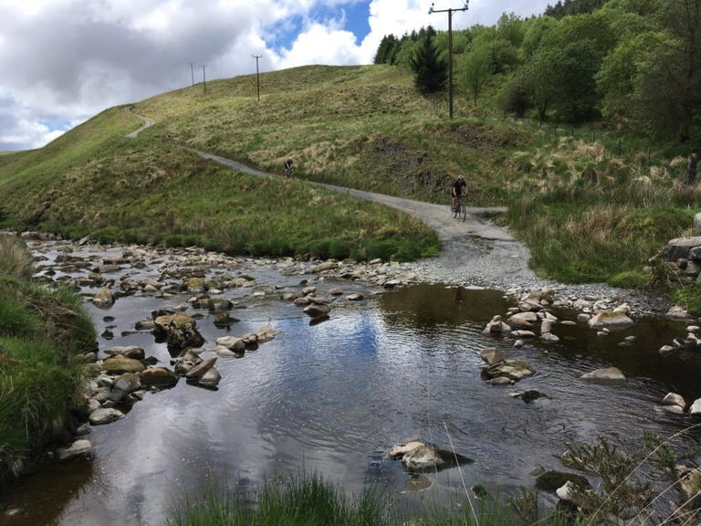Riders descend a narrow hillside lane to a rocky stream crossing on the TINAT 400km Audax route in Wales.