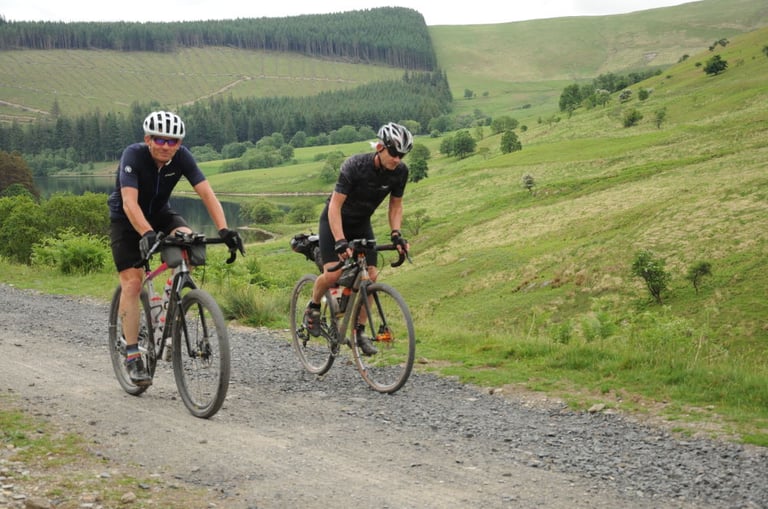 Two riders on gravel bikes climb a stony track above a reservoir during the TINAT 400km Audax in mid-Wales.