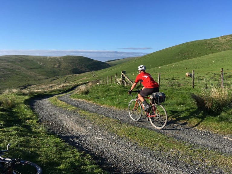 Rider in red with a saddle bag rolls down a gravel track on NCR82 through green Welsh hills under a clear sky.
