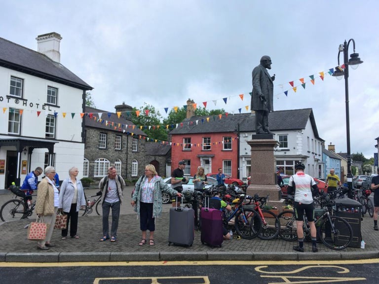 Audax riders and loaded bikes gather round a statue at the Tregaron control, watched by holidaymakers with suitcases.