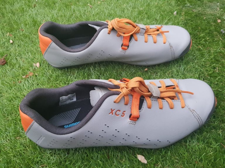Pair of Shimano XC5 shoes side-on on grass, orange laces threaded through the lace tidy and Power Strap.