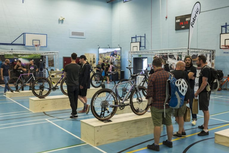 Visitors look over custom gravel bikes on wooden plinths at the Grinduro Custom Bike Expo in a school sports hall.