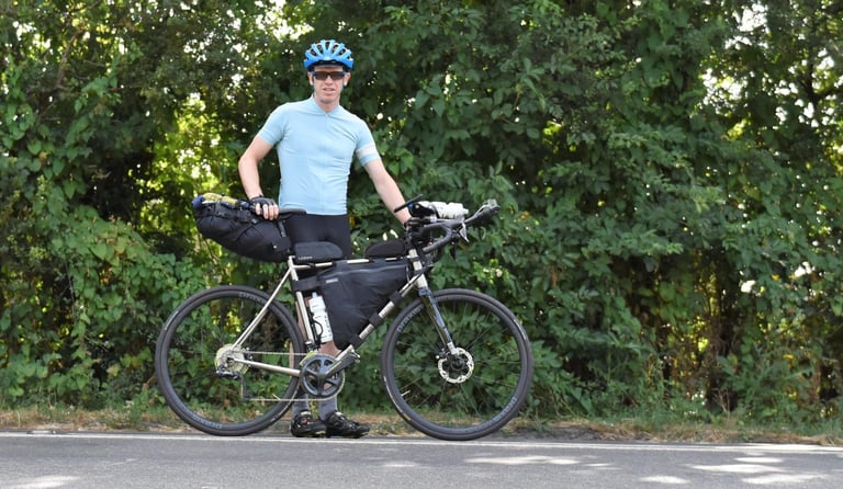 Rider stands beside a Kinesis GTD titanium bike loaded with bikepacking bags on a country road.