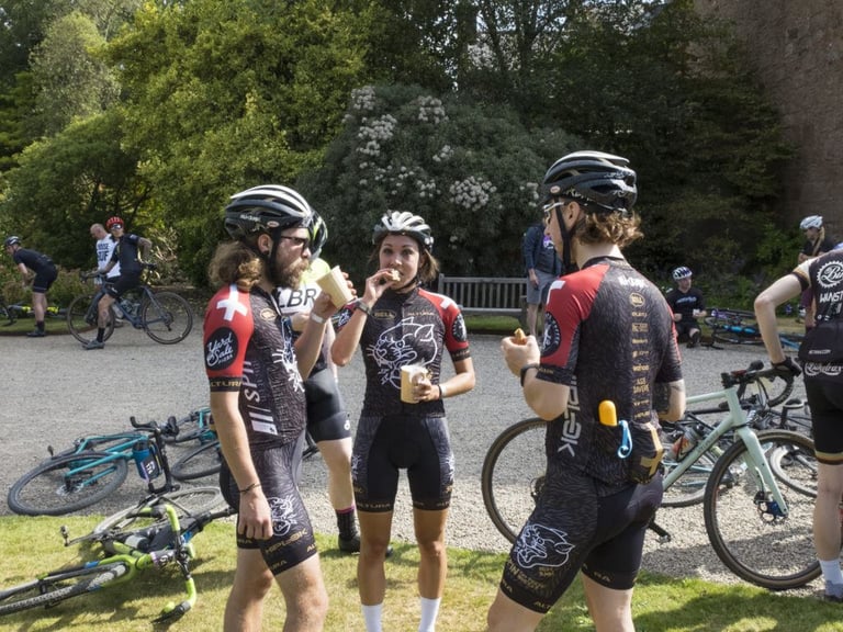 Three riders in matching team kit eat and chat in a garden at a Grinduro feed station, bikes lying on the gravel.