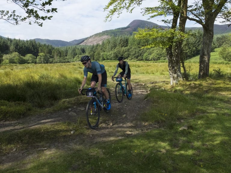 Two riders on gravel bikes pedal along a rocky track through a green valley with Arran's hills behind them.