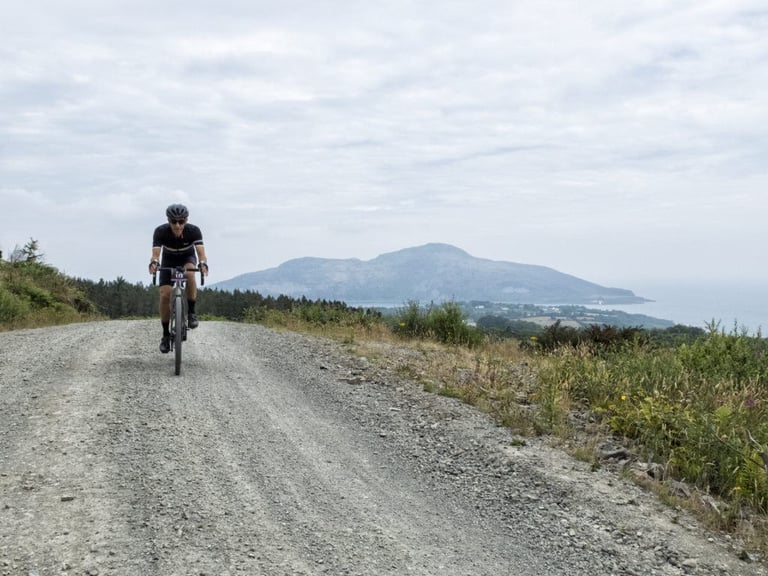 Grinduro rider on a wide gravel road on Arran, with a hilly island and the sea in the distance.