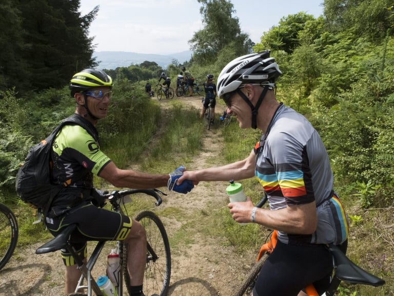 Two Grinduro riders shake hands on a grassy forest track while others ride along the path behind them.