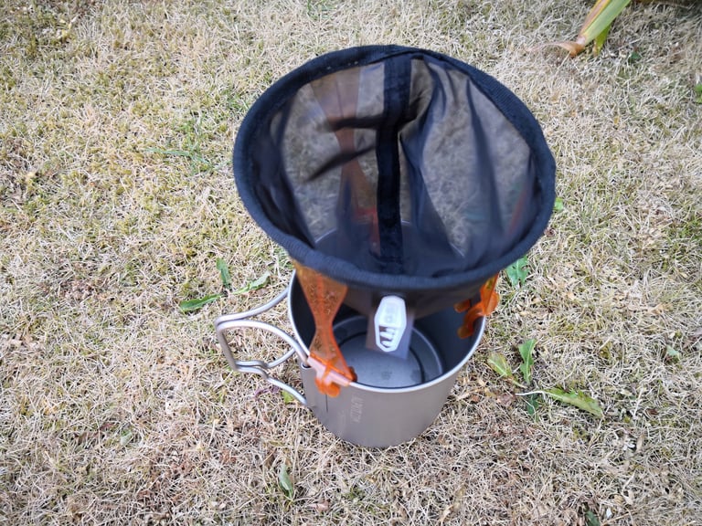 GSI Outdoors Java Drip mesh filter with orange legs clipped on the rim of a titanium mug, seen from above.