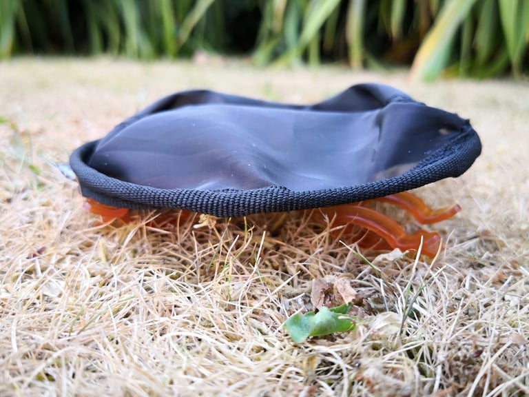 GSI Java Drip folded flat on dry grass, the black mesh filter collapsed over its orange legs, side view.