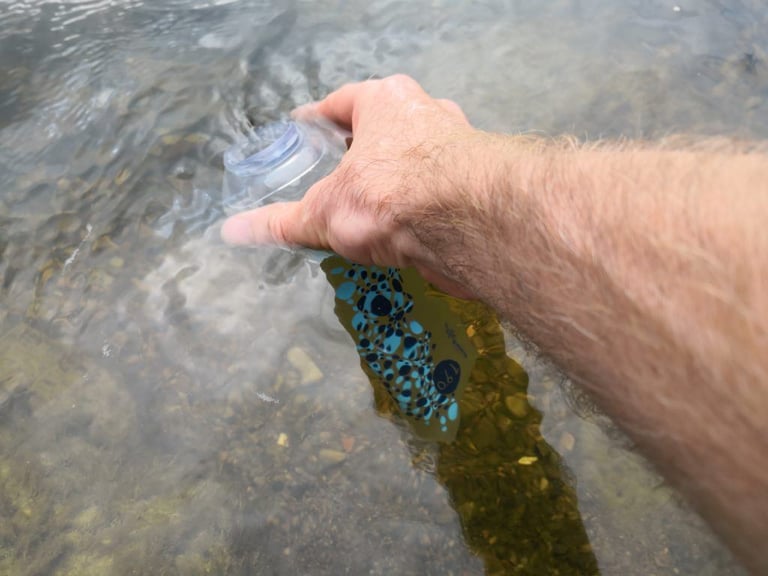 Hand holding a Katadyn BeFree soft flask under the surface of a clear stream to fill it with water.