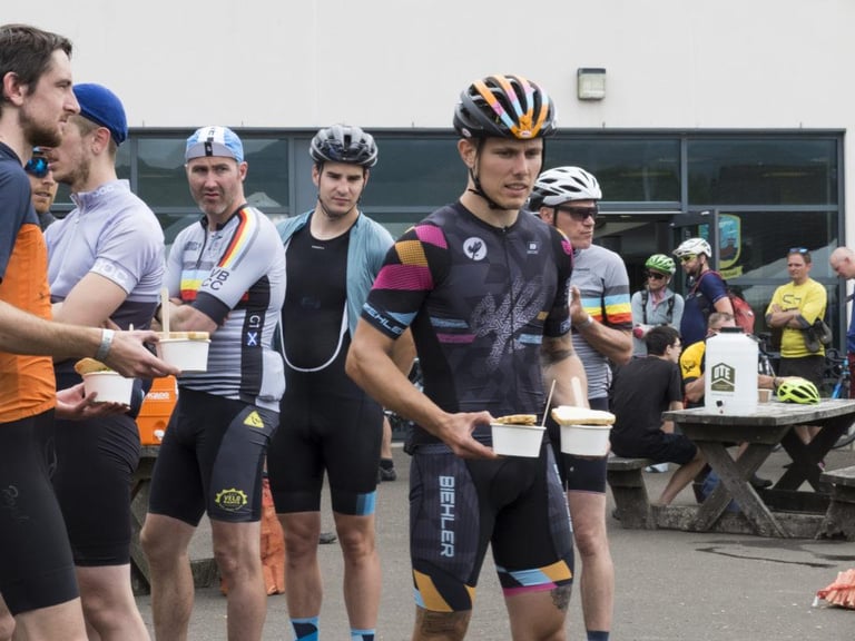 Riders in cycling kit stand outside the school holding bowls of food at the Grinduro lunch stop on Arran.