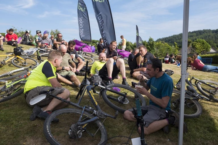 Riders relax on the grass among bikes and banners after racing Grinduro Scotland, with wooded hills behind.