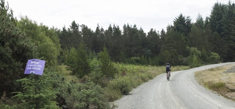 Rider on a gravel forest road at Grinduro Arran passes a purple sign reading "Smile! It makes you look faster".