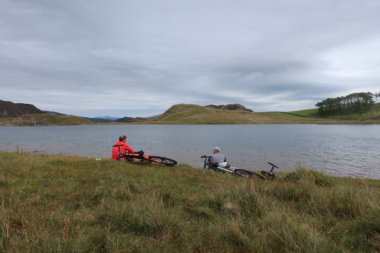 Two riders sit on the grass beside a still lake, bikes laid down, on a guided Bike Corris weekend in Wales.