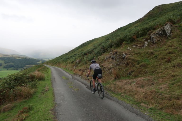 Rider on a gravel bike climbs a single-track lane below a bracken and rock hillside, with a misty valley behind, in Wales.