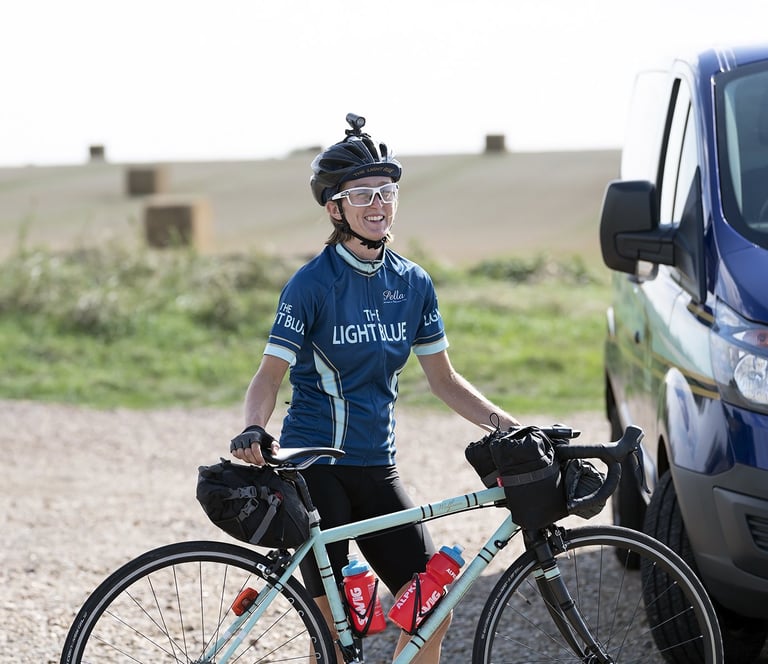 Ede Harrison smiling in a Light Blue jersey as she holds her loaded Wolfson beside the team van.