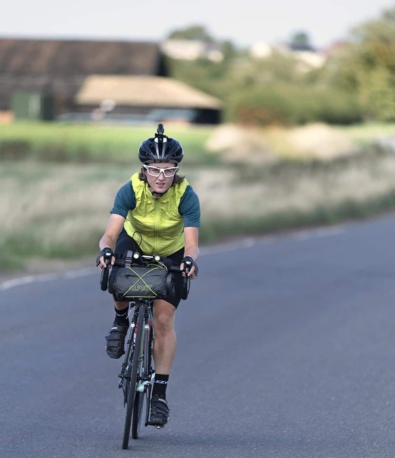 Ede Harrison rides towards the camera on a quiet road with an Alpkit handlebar bag, helmet light and white glasses.