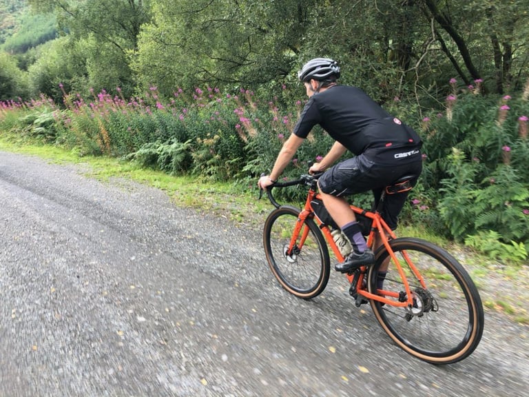 Rider on an orange gravel bike with a frame bag rides a Dyfi Forest gravel road lined with rosebay willowherb.