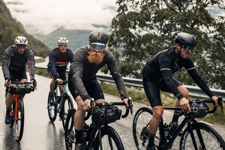 Four riders with Apidura handlebar bags ride a wet road below cloud-covered mountains on a Velochef ride.