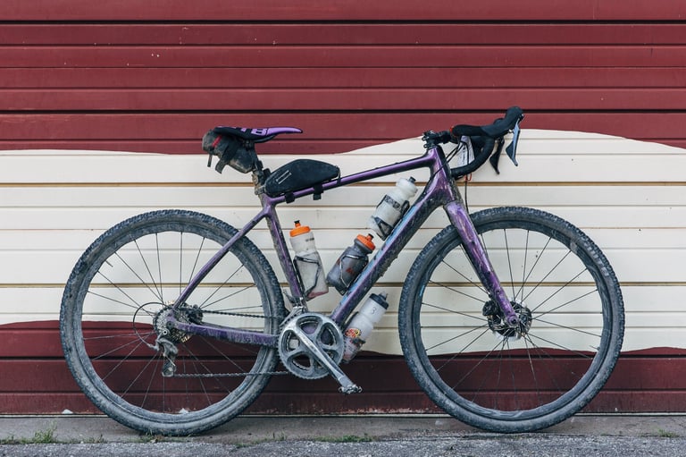 Mud-spattered purple Kona Libre with bottles and frame bag leaning on a wall after 200 miles of Dirty Kanza.