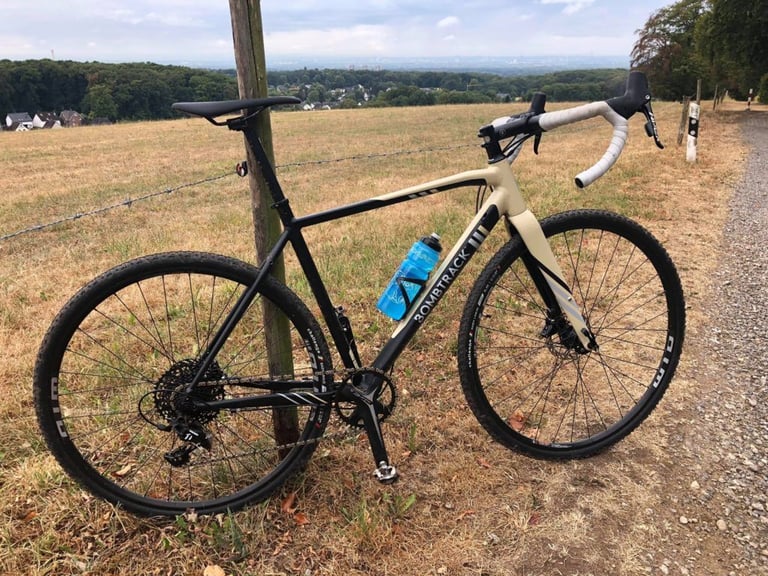 Bombtrack Tension 1 cyclocross bike in cream and black with white bar tape, beside a gravel track above a valley.