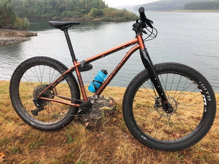 Copper-coloured Bombtrack Beyond+ 2 with fat 27.5+ tyres and a rigid carbon fork beside a reservoir.