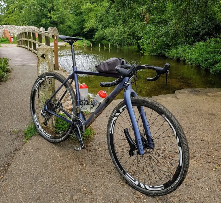 Muddy Lauf True Grit with a top tube bag and two bottles beside a river ford and footbridge on a Peddars Way tour.