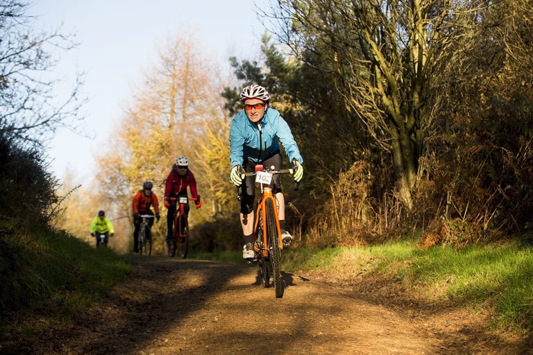 Top of the Rock gravel sportive riders, led by one on an orange bike with a race number, on a sunlit woodland trail.
