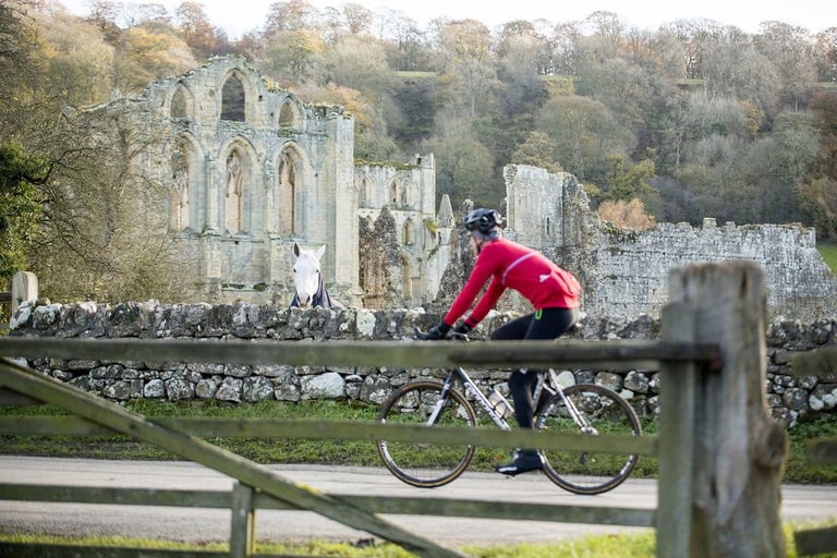 Rider in a red jacket passes a ruined abbey in North Yorkshire as a white horse looks over the wall, autumn trees behind.