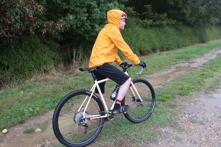 Side view of a rider pedalling with the MTR Shell Jacket's hood up over a helmet on a wet grass track.