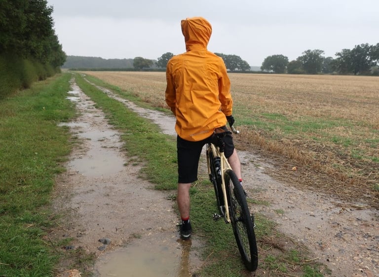 Rider with the MTR Shell's hood up stands astride a gravel bike on a puddled track beside a Suffolk stubble field.
