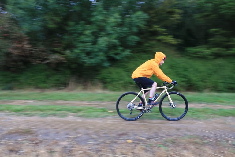 Rider in the hooded MTR Shell Jacket pedals a cream gravel bike along a wet grass track in heavy rain.