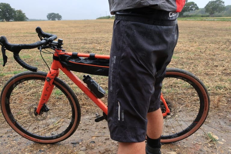 Side view of the dark grey Endura MTR Waterproof Shorts beside an orange gravel bike in a stubble field.