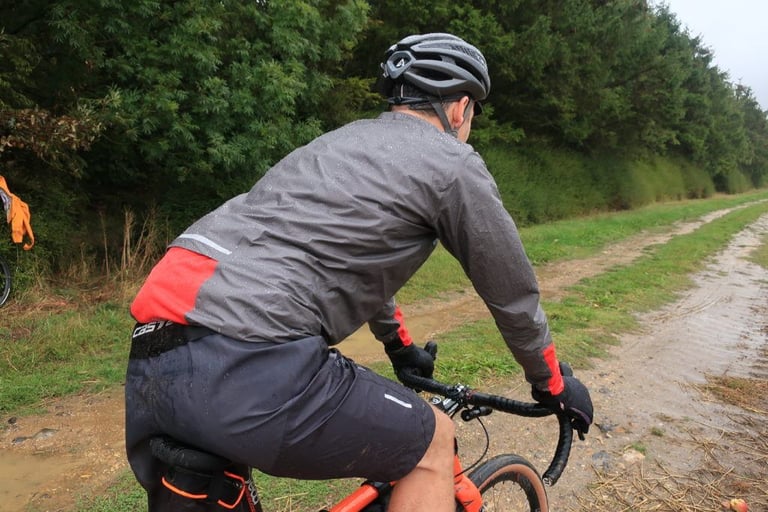 Rider in Endura MTR Waterproof Shorts and a rain-beaded grey jacket pedals along a wet grass track beside a hedge.