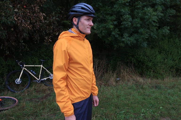 Rider wearing the Endura MTR Shell Jacket with the hood stowed in the collar, a cream gravel bike propped behind.