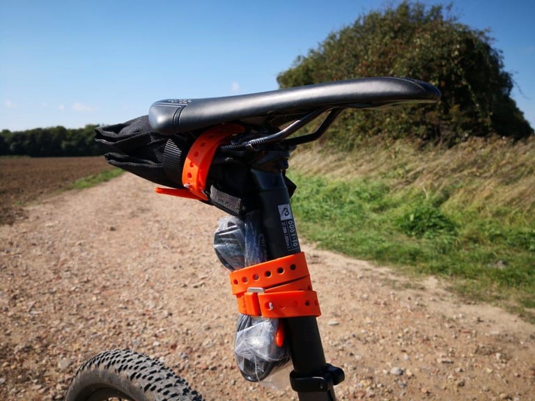 Orange Voile strap holding a tool roll under a saddle, with a spare tube strapped to the seatpost on a gravel track.