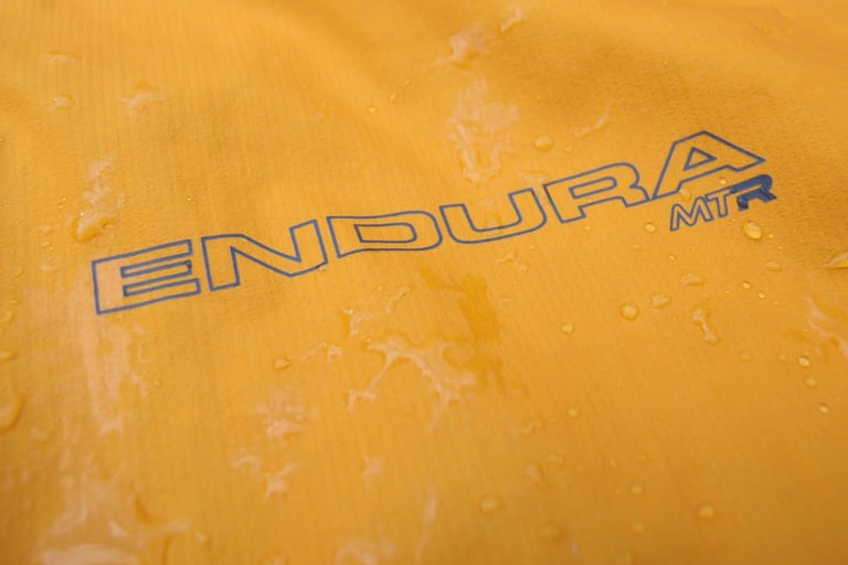 Close-up of the Endura MTR logo on the jacket's wet orange fabric, with rain beading on the surface.