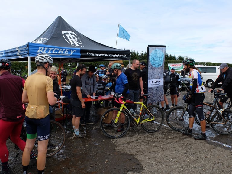 Riders gather around a Ritchey tent and a Lezyne banner at the Dirty Boar expo on a wet car park.
