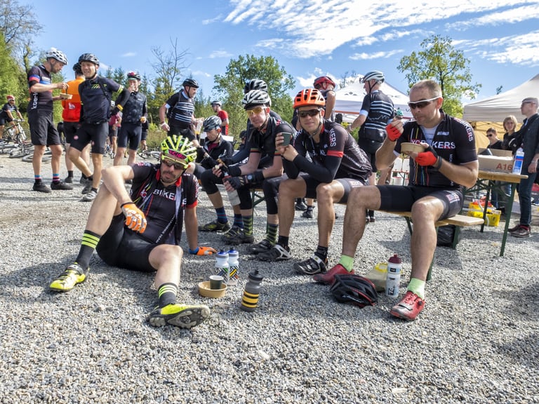 Group of riders sit on gravel and benches with bottles and bowls at a Dirty Boar rest stop in Belgium.