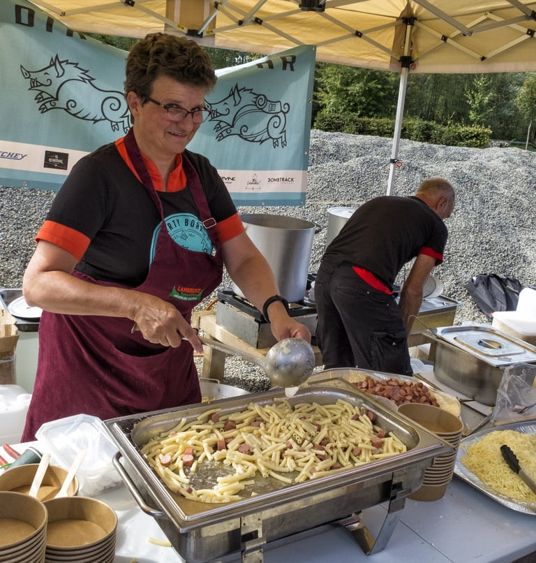 Volunteer serves macaroni and sausage from a tray at a Dirty Boar feed stop under a boar-print banner.