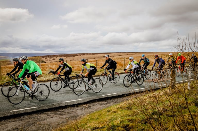 A group of road cyclists rides in a line along a tarmac road across open moorland under a cloudy sky.