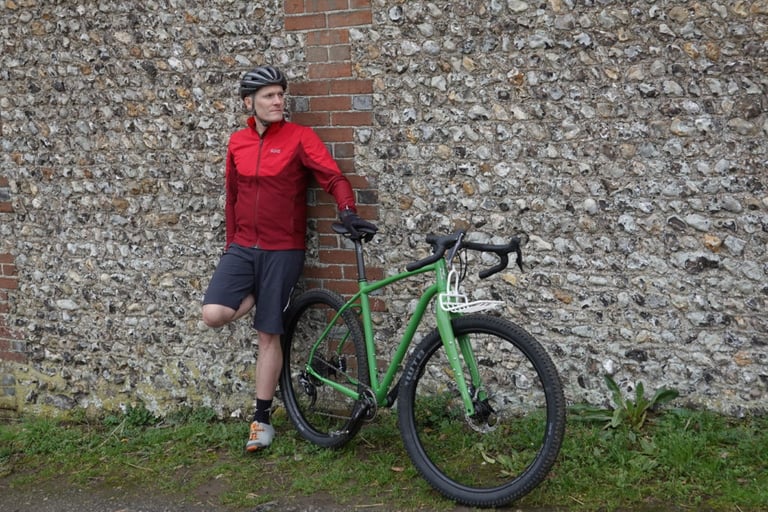 Rider wearing the red GORE R5 WINDSTOPPER shirt leans against a flint and brick wall beside a green bike.