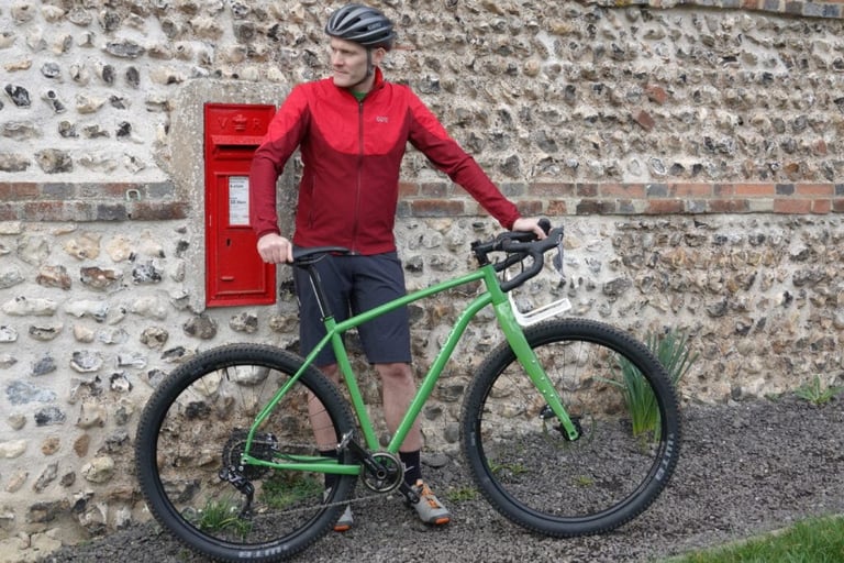 Rider in the red GORE R5 shirt stands behind a green bike next to a red Victorian postbox in a flint wall.
