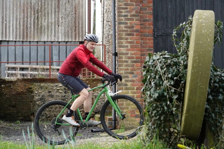 Side view of a rider in the red GORE R5 shirt pedalling a green bike past a brick farm building.